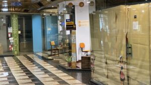Indoor shopping arcade with glass storefronts, a small orange swivel chair, and wooden seating near a information kiosk; patterned tiled floor.