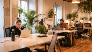Two people work at separate desks in a bright, plant-filled coworking space with laptops open and pendant lights overhead.
