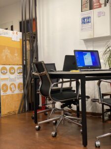 Office desk with an open laptop and black swivel chair; orange luggage storage poster on the left.
