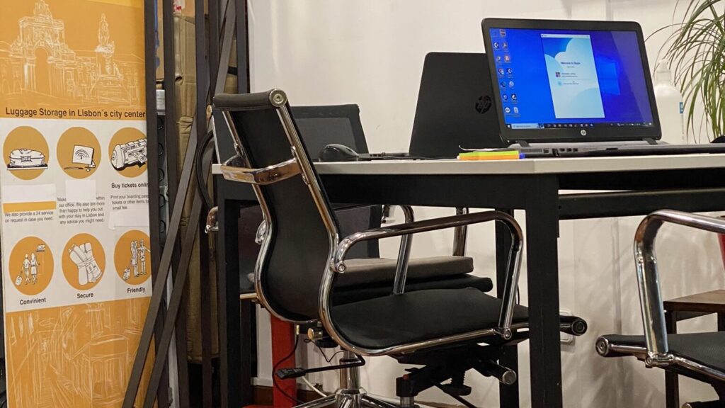 Office desk with an open laptop and black swivel chair; orange luggage storage poster on the left.