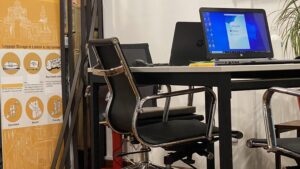Office desk with an open laptop and black swivel chair; orange luggage storage poster on the left.