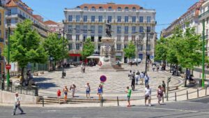 Public square with a central monument on a pedestal, surrounded by historic buildings, trees, and pedestrians on a sunny day.