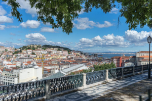Overlook of a seaside city with red-tiled rooftops, a distant river, and a decorative iron railing framing a sunny, cloud-filled sky above. Tree branches hang down from the top edge.