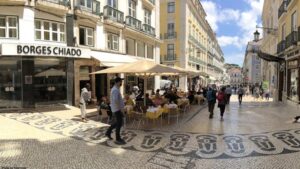 C European city street with outdoor café seating under large umbrellas, pastel buildings, and patterned stone pavement.